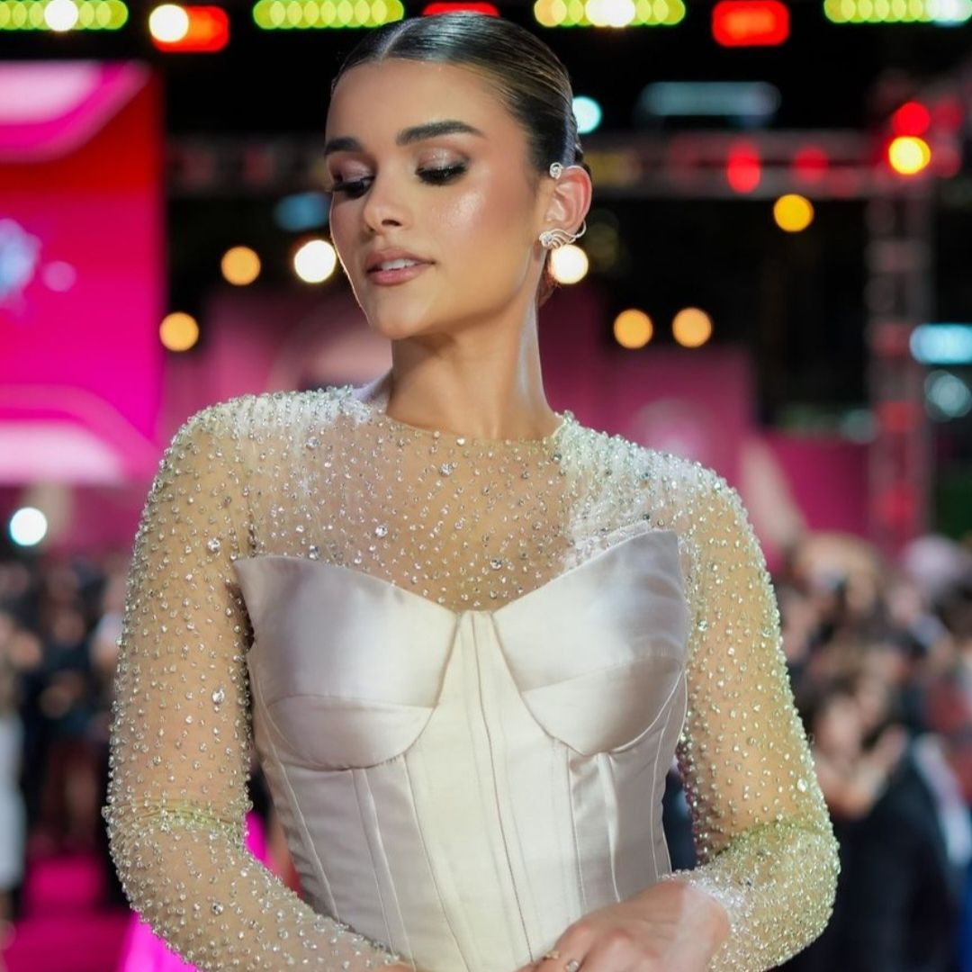 Woman in a long-sleeve ivory gown covered in rhinestones, wearing shiny earrings and glamorous makeup.