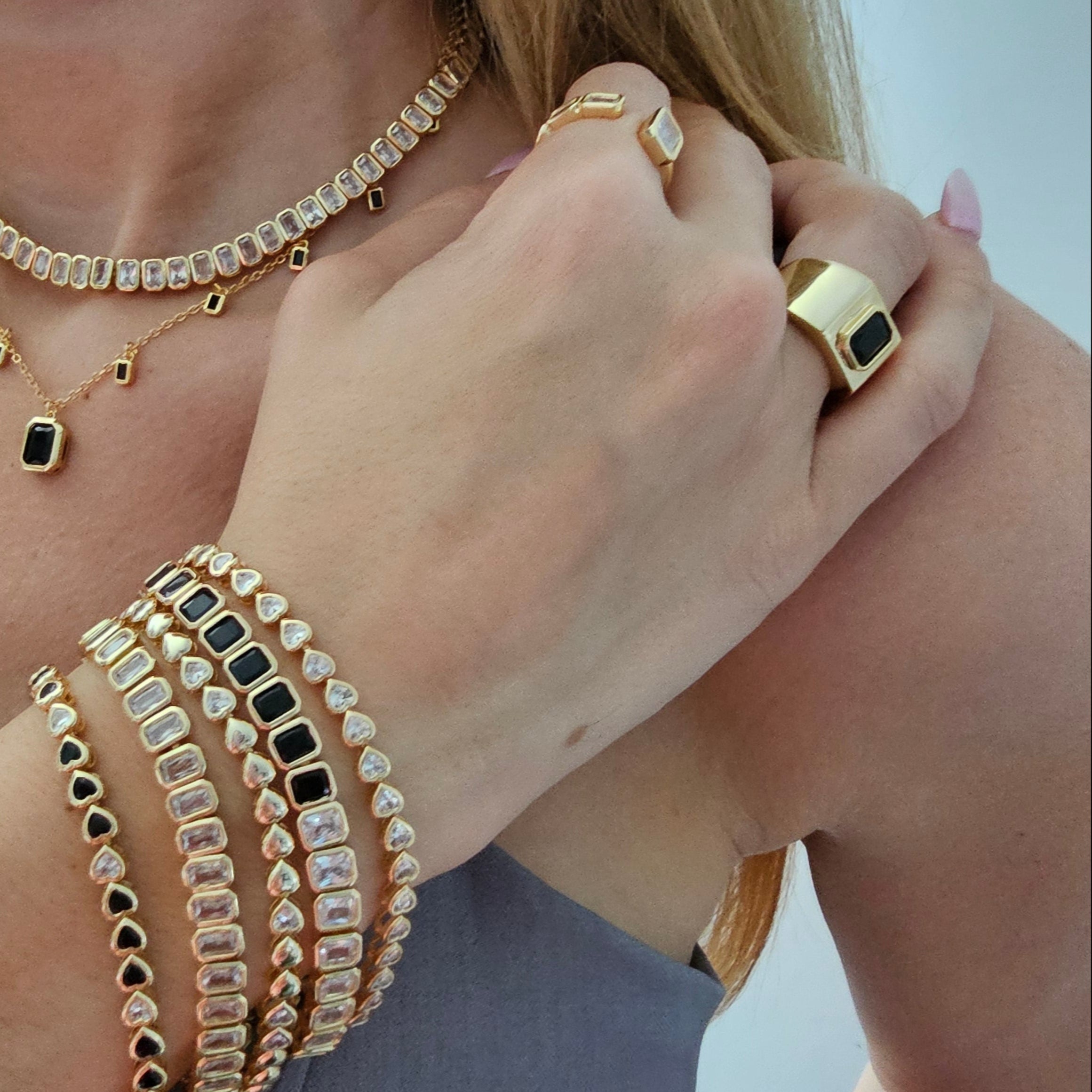 Close-up of a woman's arm wearing stacked gold bracelets with black and clear stones, layered gold necklaces with black stone pendants, and bold gold rings.