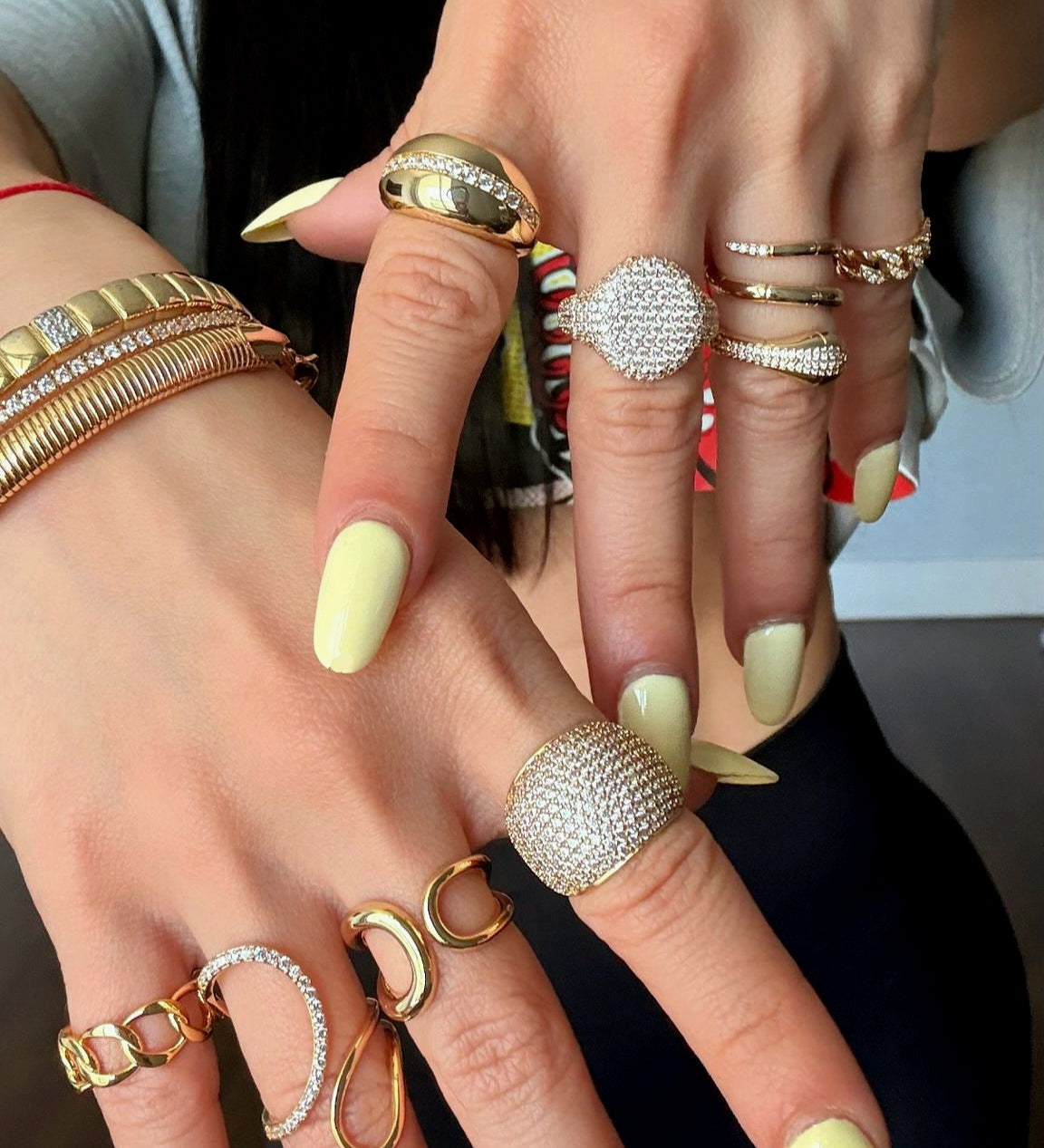 Close-up of two hands wearing multiple gold rings with pavé crystals and bold designs, paired with gold bracelets, styled with pastel yellow manicure.