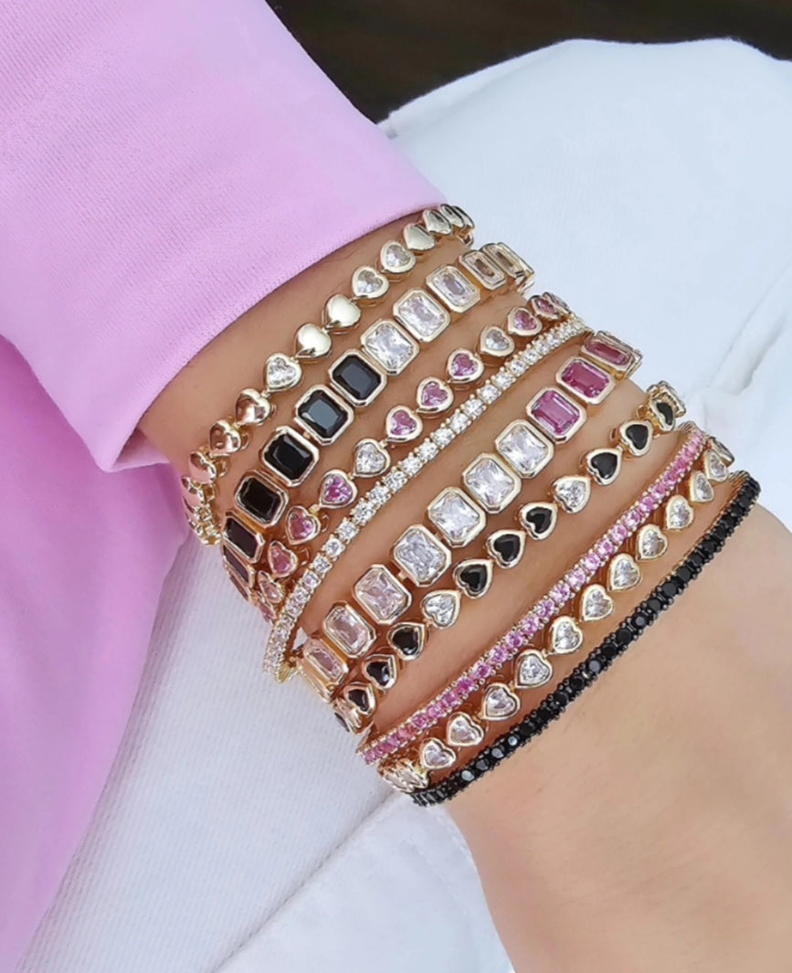 Close-up of a woman's wrist stacked with multiple gold bracelets featuring heart-shaped charms and rectangular stones in black, pink, and clear designs, paired with sparkling rhinestone accents.