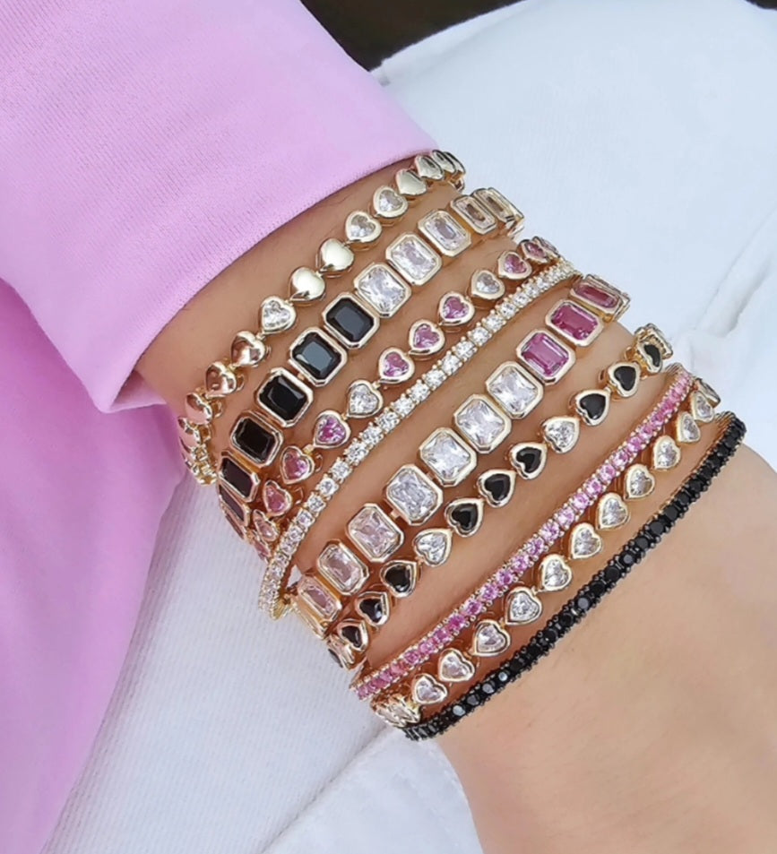 Close-up of a woman's wrist stacked with multiple gold bracelets featuring heart-shaped charms and rectangular stones in black, pink, and clear designs, paired with sparkling rhinestone accents.