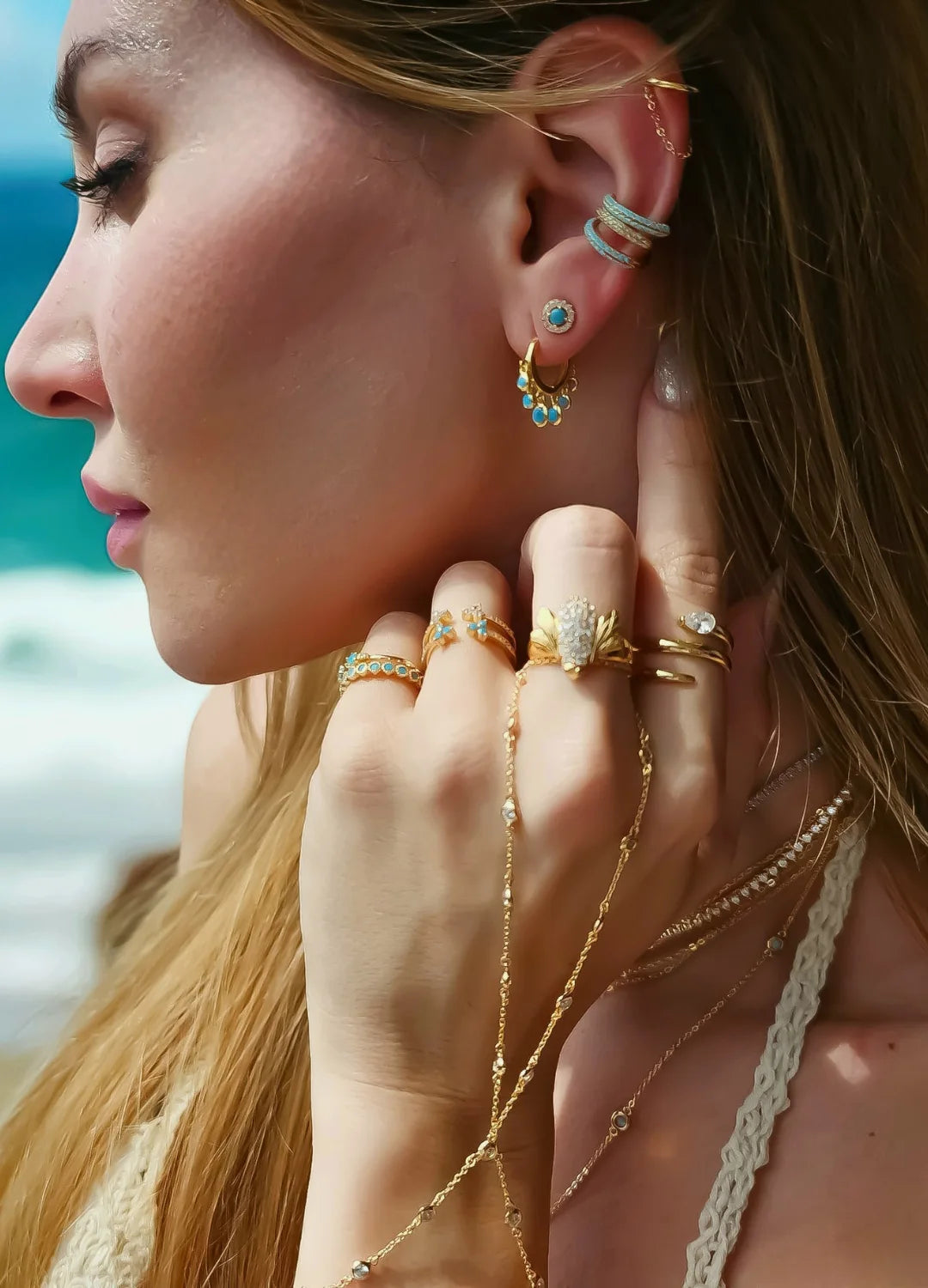Woman wearing elegant earrings and rings, standing by the beach with the ocean in the background. The jewelry reflects the sunlight, highlighting its fine design and craftsmanship.