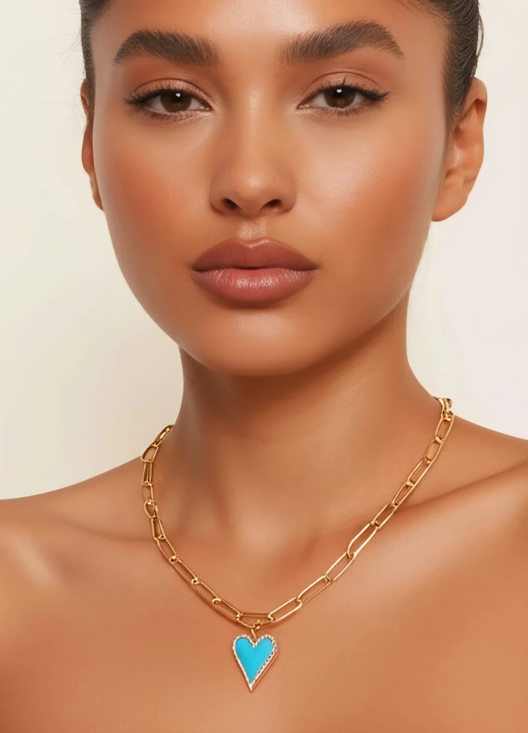 Woman wearing a heart-shaped necklace against a soft light background. The focus is on the elegant, minimalist design of the pendant, highlighting its delicate curves and romantic symbolism.