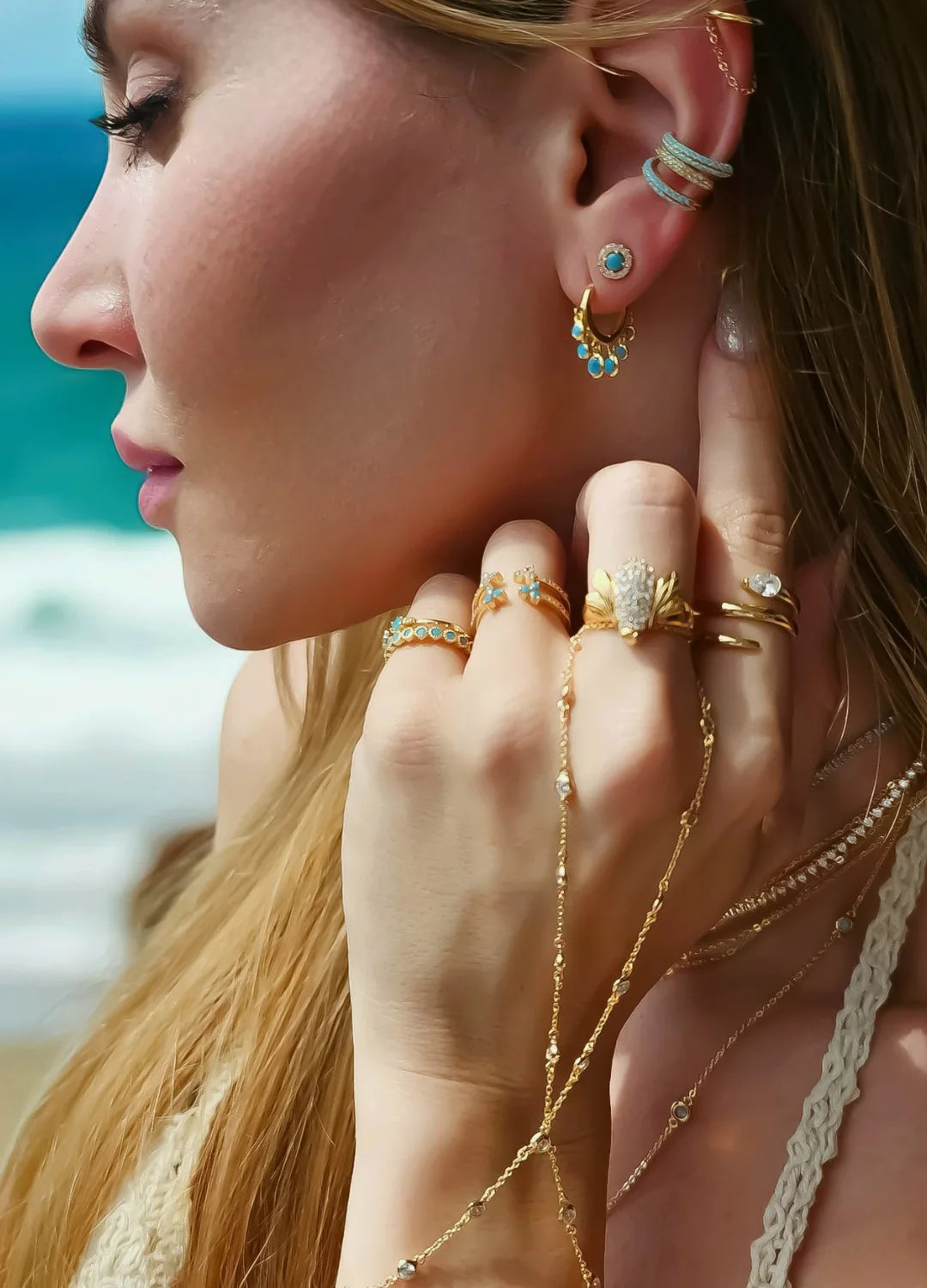 Woman wearing elegant earrings and rings, standing by the beach with the ocean in the background. The jewelry reflects the sunlight, highlighting its fine design and craftsmanship.